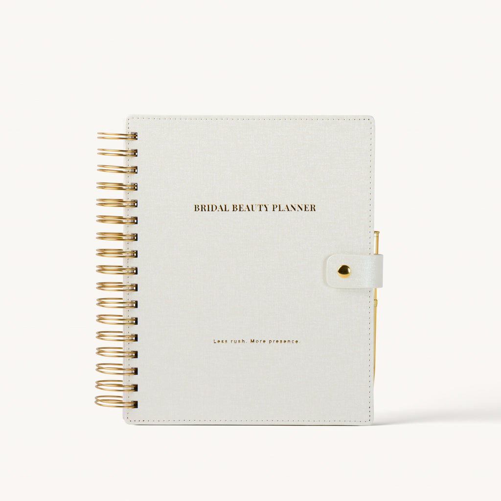 A bridal beauty planner notebook with a textured off-white cover, gold wire spiral binding on the left, and a gold snap closure on the right. The cover features the text 'BRIDAL BEAUTY PLANNER' in gold.