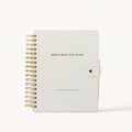A bridal beauty planner notebook with a textured off-white cover, gold wire spiral binding on the left, and a gold snap closure on the right. The cover features the text 'BRIDAL BEAUTY PLANNER' in gold.