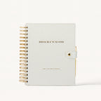 A bridal beauty planner notebook with a textured off-white cover, gold wire spiral binding on the left, and a gold snap closure on the right. The cover features the text 'BRIDAL BEAUTY PLANNER' in gold.