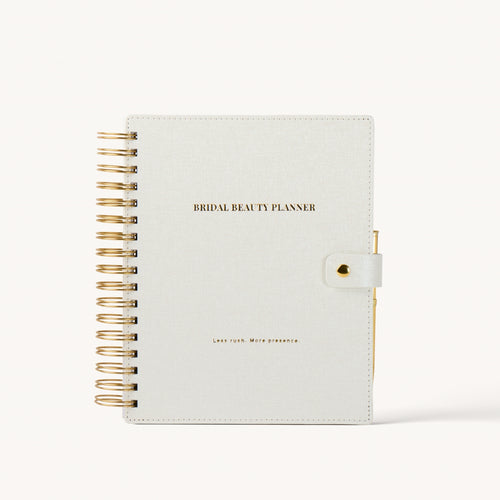 A bridal beauty planner notebook with a textured off-white cover, gold wire spiral binding on the left, and a gold snap closure on the right. The cover features the text 'BRIDAL BEAUTY PLANNER' in gold.
